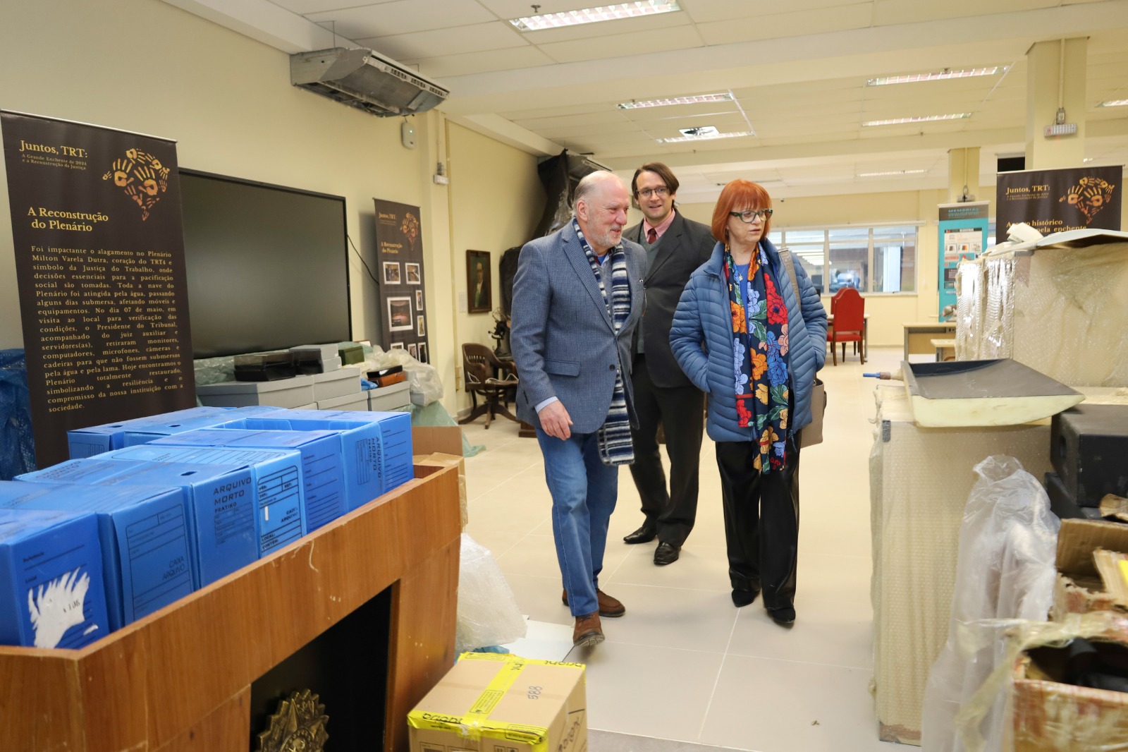 Três pessoas andam em direção ao canto direito da imagem. O ambiente é a sala do memorial. Diversos materiais estão embalados em caixas e sacos plásticos.